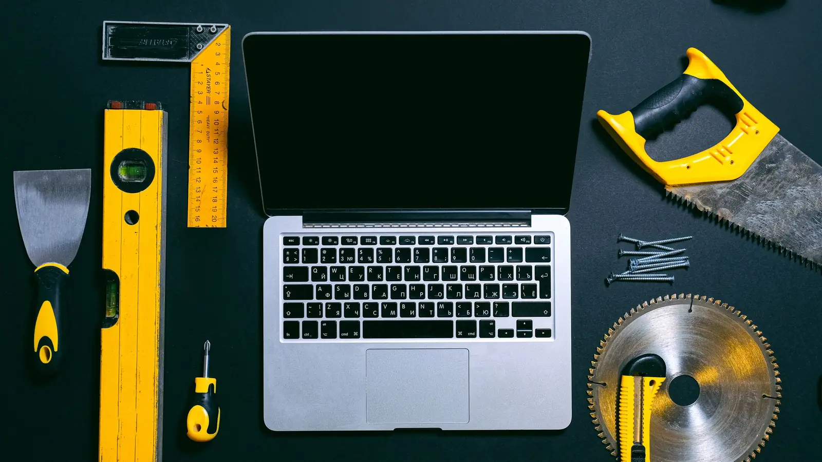 Laptop pe un fundal închis, înconjurat de unelte: nivelă, echer, fierăstrău și șurubelniță
